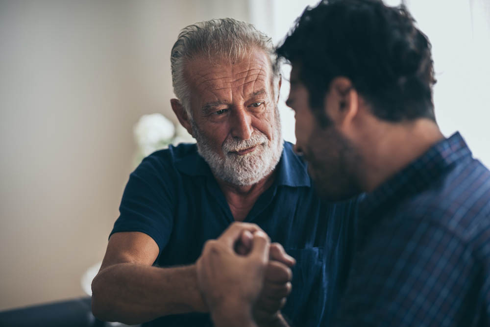Sweet bonding. Cheerful elderly man sitting on the sofa next to his adult son, hugging him and posing together with him
