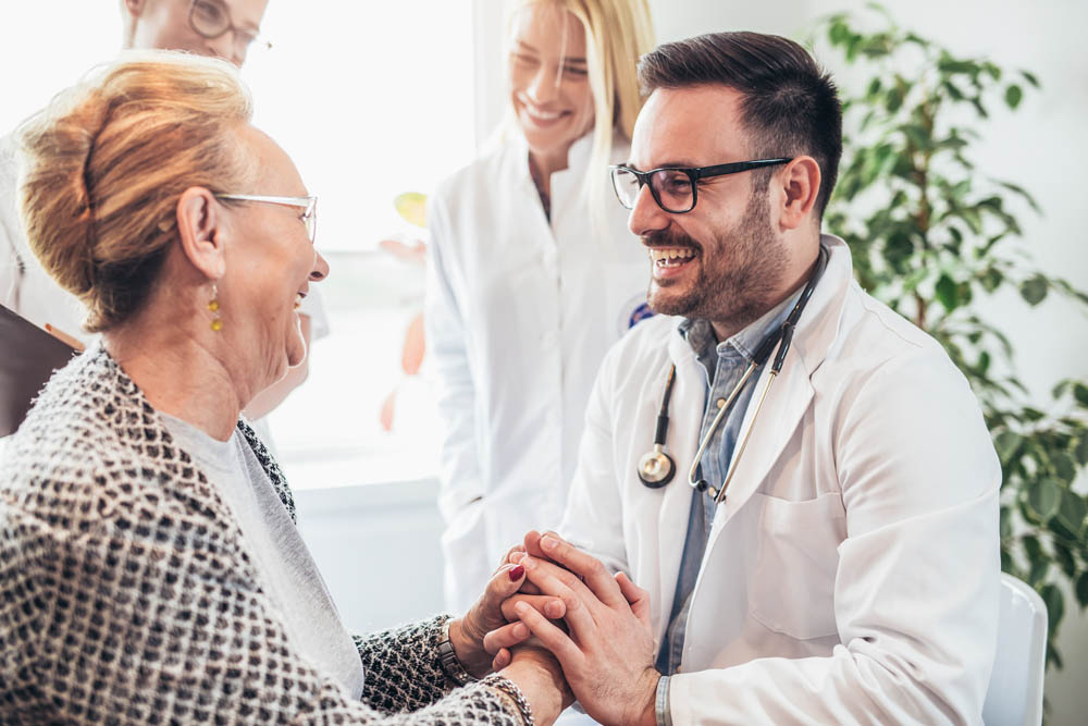 An elderly woman smiling and holding hands with a doctor in a medical setting.