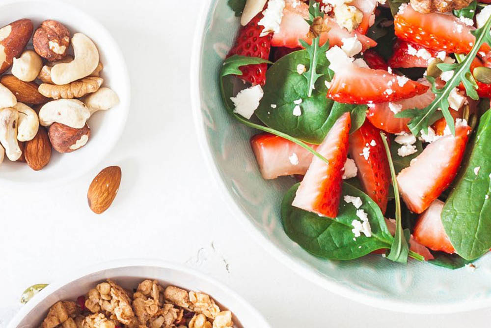 Close-up of a salad with strawberries, spinach, and cheese in a bowl, accompanied by bowls of mixed nuts and granola.